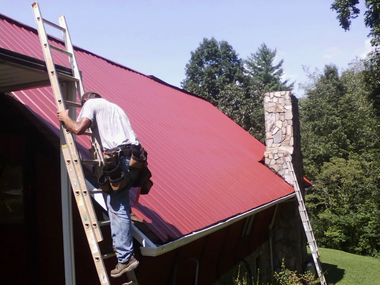 Roofer on a ladder beside a red metal roof and stone chimney — Soffit & Fascia Repair by Roofing Brothers in Wilkesville